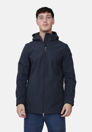 Giovane uomo che indossa una giacca con cappuccio blu scuro, con la cerniera chiusa, in piedi davanti a uno sfondo chiaro semplice, con le mani rilassate lungo i fianchi.