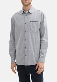 Chemise grise à manches longues avec un motif texturé, devant boutonné, boutons marron et une poche poitrine unique avec un accent bleu.