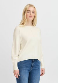 Sweter o kremowym kolorze, dziany, luźny, z okrągłym dekoltem i długimi rękawami. Posiada szew na przodzie oraz ściągacze na rękawach i u dołu.