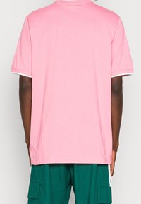 Persona che indossa una maglietta rosa a maniche corte larga con rifiniture bianche sulle maniche e pantaloni cargo verde scuro con tasche laterali, mostrata di spalle.