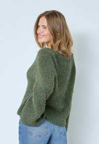 Zaļš, fakturēts adījuma džemperis ar brīvu piegriezumu, ar garām piedurknēm un apaļu kakla izgriezumu, kombinēts ar ziliem džinsiem.