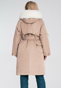 Beżowy długi płaszcz z sztucznym futrem przy kołnierzu, z paskiem w talii, dużymi kieszeniami i luźnym krojem. Gładka tekstura i praktyczny design.