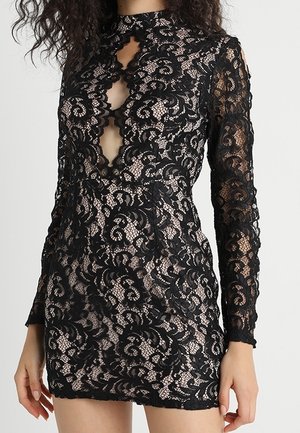 Nainen pukeutuneena vartalonmyötäiseen, pitkähihaiseen mustaan pitsiseen minimekkoon, jossa on ihonsävyinen vuori ja avonainen pääntie.