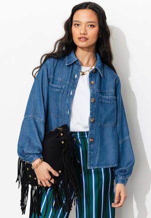 Femme portant une veste en jean bleue sur une chemise blanche, un pantalon rayé multicolore, tenant un sac noir à franges avec des clous métalliques.