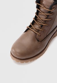 Botas de invierno de cuero marrón con cordones beige, forro de felpa negra y suela de goma resistente sobre un fondo blanco.