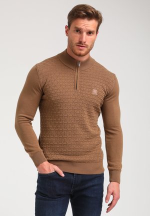 Bruine gebreide pullover met een halve rits kraag, voorzien van een geperforeerd geometrisch patroon en geribbelde manchetten. Draagt met donkere jeans.