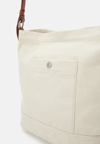 Borsa a spalla in tela beige con una tracolla in pelle, presenta una tasca frontale con un bottone a pressione argento, e un design resistente e testurizzato.