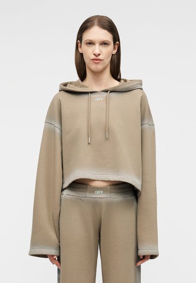 Γυναίκα με φαρδιά μπεζ cropped φούτερ με κουκούλα και ταιριαστό παντελόνι ψηλόμεσο με το λογότυπο "OFF", στέκεται μπροστά σε απλό ανοιχτό φόντο.