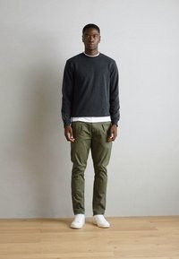 Cămașă lungă gri închis, cu un tricou alb pe dedesubt, pantaloni verzi și adidași albi; stând pe un podea din lemn deschis, în fața unui perete gri.