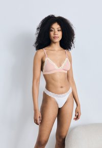 Młoda kobieta o kręconych włosach, ubrana w różowy biustonosz z koronkową lamówką i białe koronkowe majtki, stojąca na tle jednolitego jasnoszarego tła.