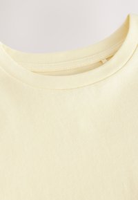 Detailní záběr na světle žluté tričko s kulatým výstřihem, zobrazující límec a horní část hrudi s viditelnými detaily šití.