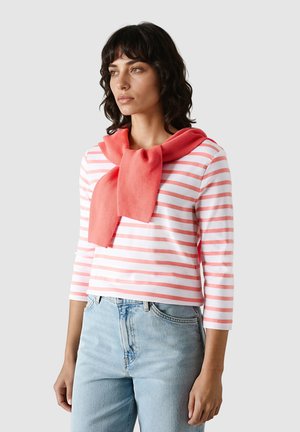 Žena s černými kudrnatými vlasy, která má na sobě růžové a bílé pruhované tričko s dlouhým rukávem, světle modré džíny a korálový svetr přehozený přes ramena.