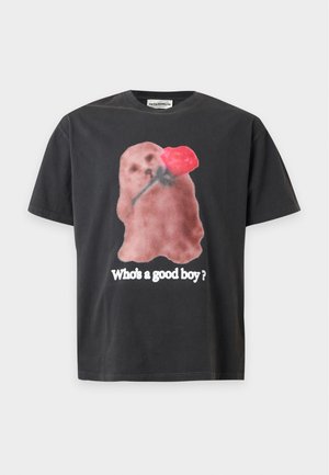 Νέος άνδρας που φοράει μαύρο T-shirt με έναν σκύλο που κρατάει μια κόκκινη τριανταφυλλιά και το κείμενο "Who's a good boy?", μαύρα σορτς μέχρι το γόνατο, λευκές κάλτσες και μαύρα παπούτσια.
