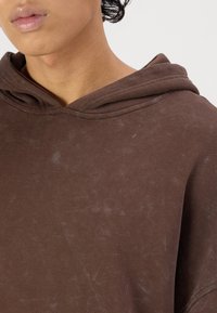 Sudadera marrón hecha de tela suave, con una gran capucha, textura visible y patrones más claros difusos en toda la prenda. Vista en primer plano.