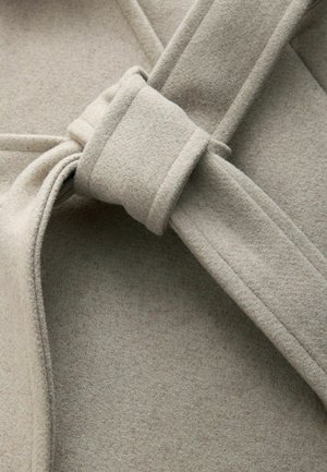 Helebeežist villasegust kangast, millel on sõlme detail ja sile tekstuur, nähtava puuvillase õmblusega ning minimalistlik, volditud disain.