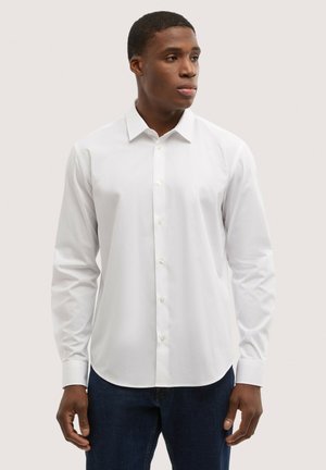 Mężczyzna w prostym białym koszuli na guziki z długim rękawem i ciemnoniebieskich dżinsach, stojący i patrzący w prawo na neutralnym tle.