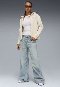 Cremfarbener Reißverschlussjacke, weißes enges Oberteil, hellblaue weit geschnittene Jeans und weiße Sneakers. Model trägt dunkle Sonnenbrille, die Frisur ist geflochten.