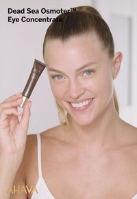 Tubetto marrone di AHAVA Dead Sea Osmoter™ Eye Concentrate con dettagli dorati, tenuto da una persona in una maglietta bianca, sorridente. Pelle liscia, sfondo chiaro.