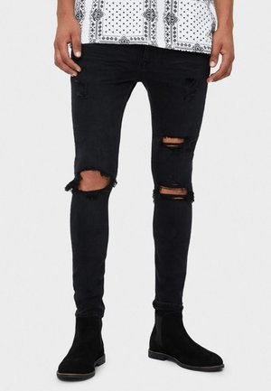 Homme portant un jean skinny noir déchiré et des bottines Chelsea en suède noir, chemise blanche à motifs partiellement visible, debout sur un fond blanc.