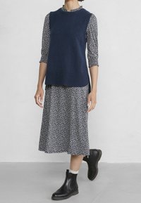 Marinblå ärmlös tröja över en lång, mönstrad klänning med vita blommor; modellen bär svarta ankelskots med vit kant. Släta material, avslappnad design.