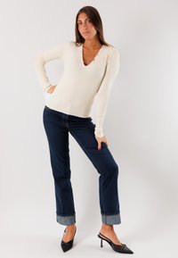 Pullover a coste color crema con scollo a V e tre applicazioni a catena. Abbinato a jeans a gamba dritta blu scuro e décolleté nere a punta.