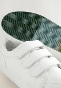 Weiße Sneaker mit einem glatten synthetischen Obermaterial, drei Klettverschlüssen und einer strukturierten grünen und weißen Gummisohle mit Fischgrätenmuster.