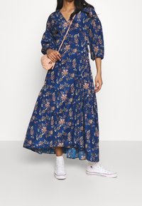 Robe maxi bleu à fleurs avec des manches bouffantes, un col en V et une jupe à volants, associée à des baskets blanches et un petit sac crossbody rose.