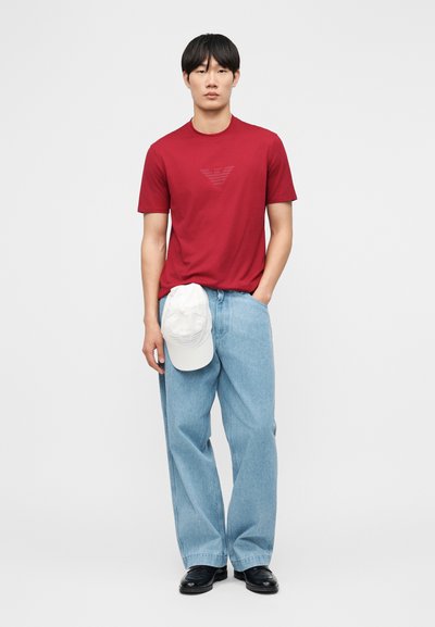 Νέος άνδρας που φορά κόκκινο μπλουζάκι, ανοιχτό μπλε παντελόνι με φαρδύ πόδι, μαύρα παπούτσια, κρατώντας ένα λευκό καπέλο στο πλάι του μπροστά σε ένα απλό φωτεινό φόντο.