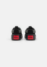 Zapatillas negras con una parte superior de malla texturada, detalles de ante y suela de goma. Logo rojo en el talón. Diseño minimalista, vista trasera.