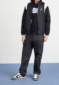 Černá bunda na zip s bílými detaily, černé tričko, černé cargo kalhoty a černé/bílé/červené tenisky s vzorovaným designem.