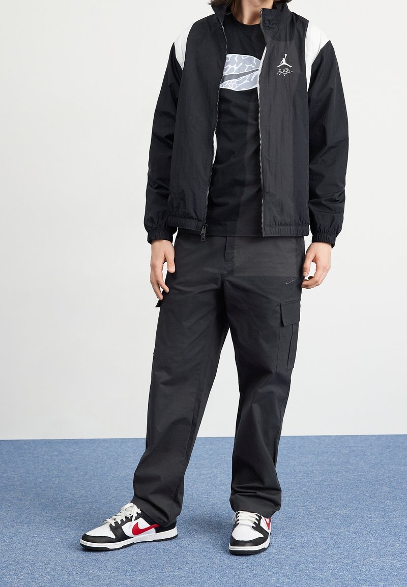 Černá bunda na zip s bílými detaily, černé tričko, černé cargo kalhoty a černé/bílé/červené tenisky s vzorovaným designem.