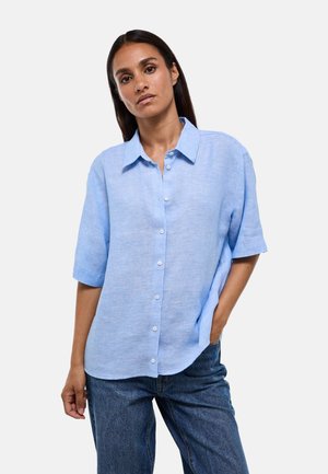 Femme aux longs cheveux foncés portant une chemise bleu clair à manches courtes boutonnée et un jean bleu, debout avec une main dans sa poche.