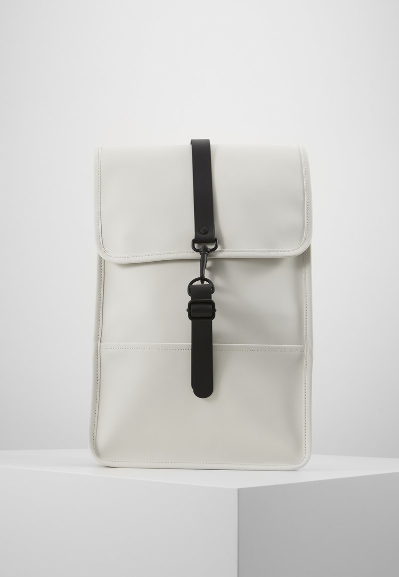 Sac à dos minimaliste gris clair avec un rabat supérieur maintenu par une sangle noire verticale avec boucle, posé sur un piédestal blanc sur fond uni.
