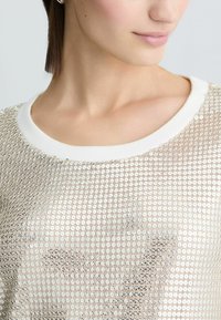 Camicetta beige chiaro con scollo bianco, caratterizzata da un motivo di piccole paillettes circolari lucide e da una consistenza morbida.