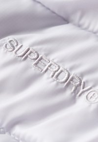 Vitt puffjacka i ett mjukt och slätt tyg; har en framträdande broderad "SUPERDRY"-logotyp i grå tråd.
