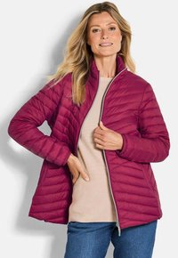 Femme aux longs cheveux blonds portant une veste matelassée rose foncé à fermeture éclair partielle sur un pull beige et un jean bleu.