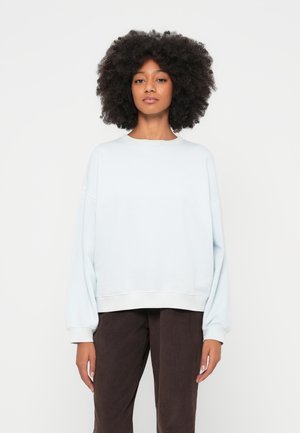 Lys blågenser med avslappet passform, senkede skuldre og ribbestrikkede mansjetter. Kombinert med mørkebrune cordbukser. Enkelt design, myk tekstur.