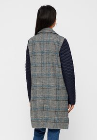 Donna con lunghi capelli scuri che indossa un cappotto scozzese di lunghezza media con maniche trapuntate blu navy, rivolta di spalle su uno sfondo chiaro e semplice.