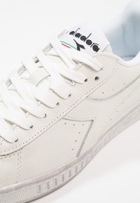 Vit lädersneaker med texturerad yta, utrustad med vita snörningar, en grå sula och en logotypad plös med gröna och röda accenter.