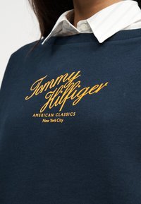 Μπλε ναυτικό πουλόβερ με κίτρινο κείμενο "Tommy Hilfiger American Classics New York City" φορεμένο πάνω από λευκό πουκάμισο με γιακά.