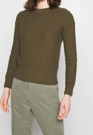 Personne portant un pull en tricot texturé vert olive et un pantalon décontracté gris clair, debout les bras détendus contre un fond uni.