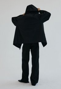 Osoba w luźnych czarnych spodniach i za dużej czarnej kurtce z kapturem, regulująca kaptur, zwrócona tyłem na jednolitym jasnym tle.