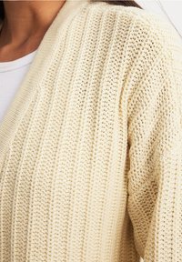 Detailní záběr na krémový chunky pletený cardigan nošený přes bílou žebrovanou košili, který zobrazuje texturované vertikální pletené vzory.