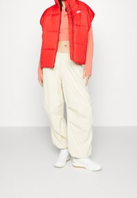 Röd vadderad ärmlös väst, rosa kort topp, krämfärgade vida byxor och vita sneakers med gumminotor; släta texturer genomgående.