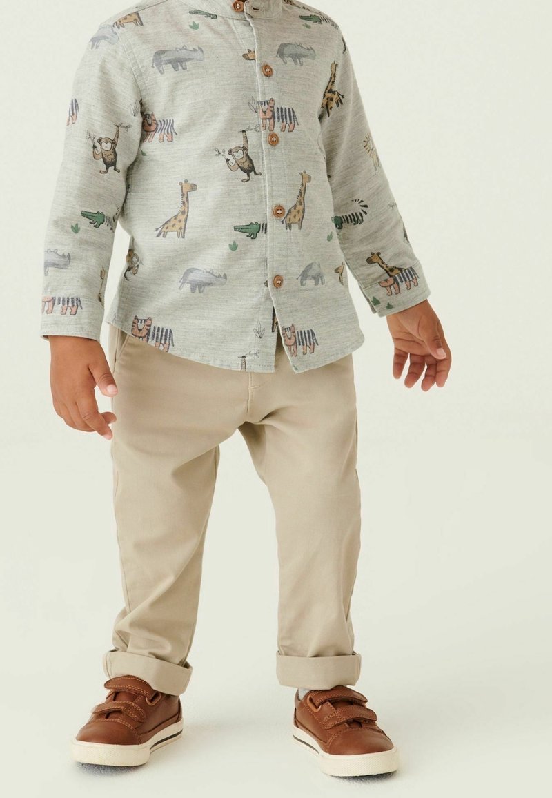 Enfant portant un pantalon beige, des baskets marron et une chemise gris clair boutonnée avec des imprimés colorés d'animaux, dont des girafes et des crocodiles.