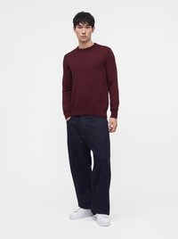 Jeune homme debout, les mains dans les poches, portant un pull bordeaux, un jean large foncé et des baskets blanches sur fond blanc.