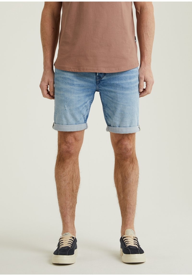 Homme portant un short en jean bleu clair retroussé, un t-shirt marron et des baskets noires avec des lacets beige, debout devant un fond uni.