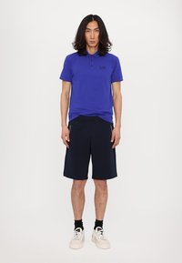 Polo blu con un logo discreto, abbinato a pantaloni corti navy e sneakers bianche. Il modello ha i capelli lunghi e si trova di fronte a uno sfondo semplice.