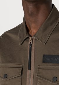 Olivengrøn jakke med lynlås foran, skulderstropper og to brystlommer med knapper. Har et mærket patch og tekstureret stof.