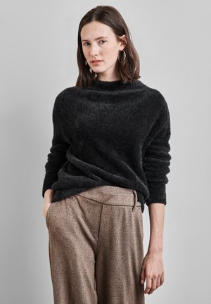 Frau mit schulterlangem braunem Haar, die goldene Creolen, einen schwarzen flauschigen Pullover und beige hochtaillierte gemusterte Hosen trägt, steht vor einer grauen Wand.